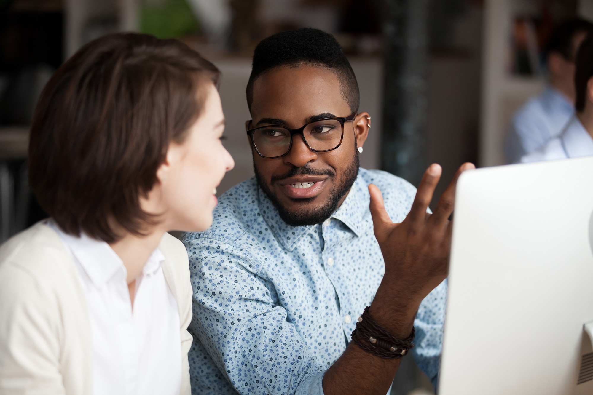 A man in glasses talks to a woman in front of a computer monitor.