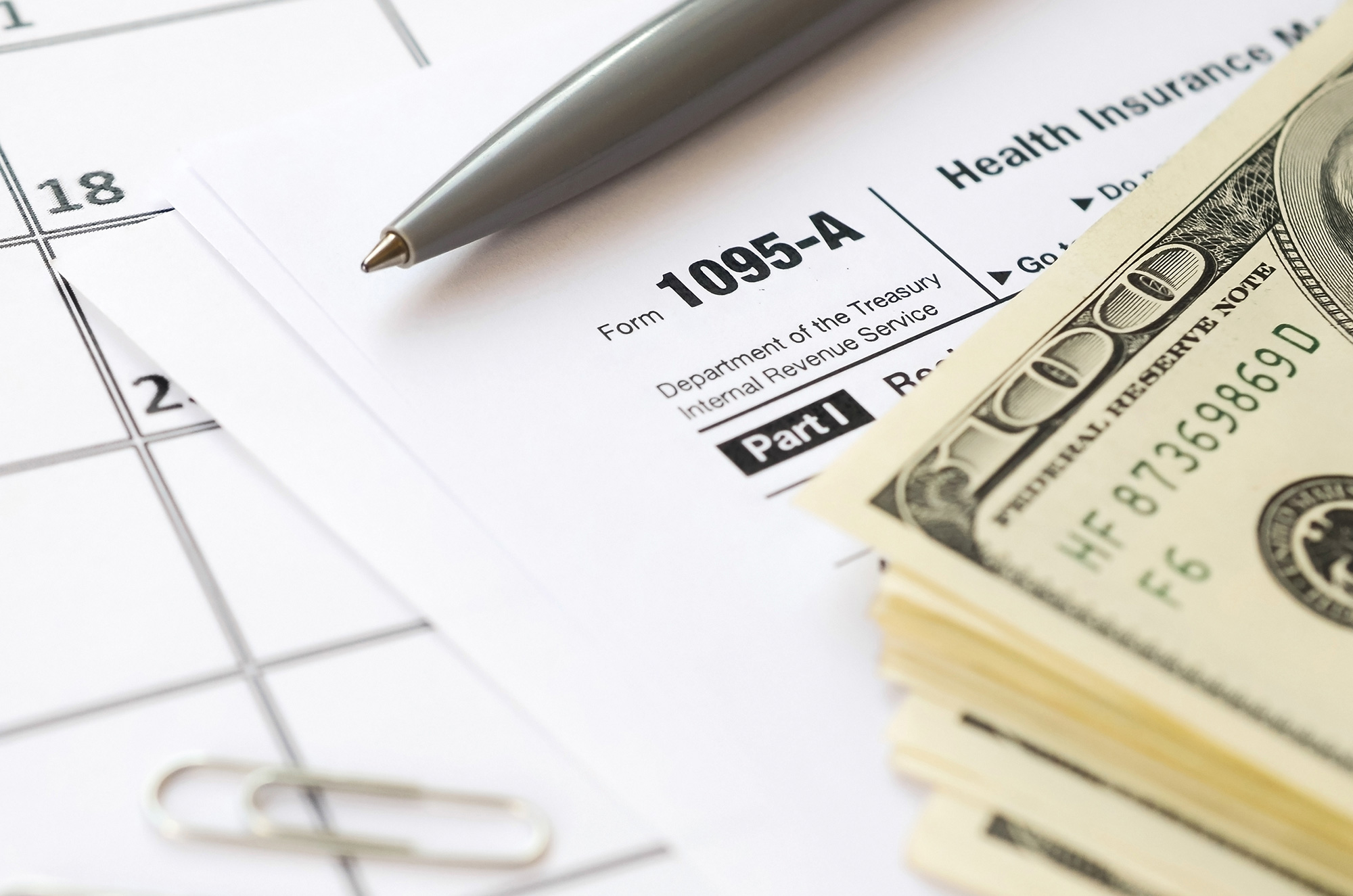 Top-down view of a calendar, a 1095 tax form, a pen and a stack of one hundred-dollar bills arranged nicely.