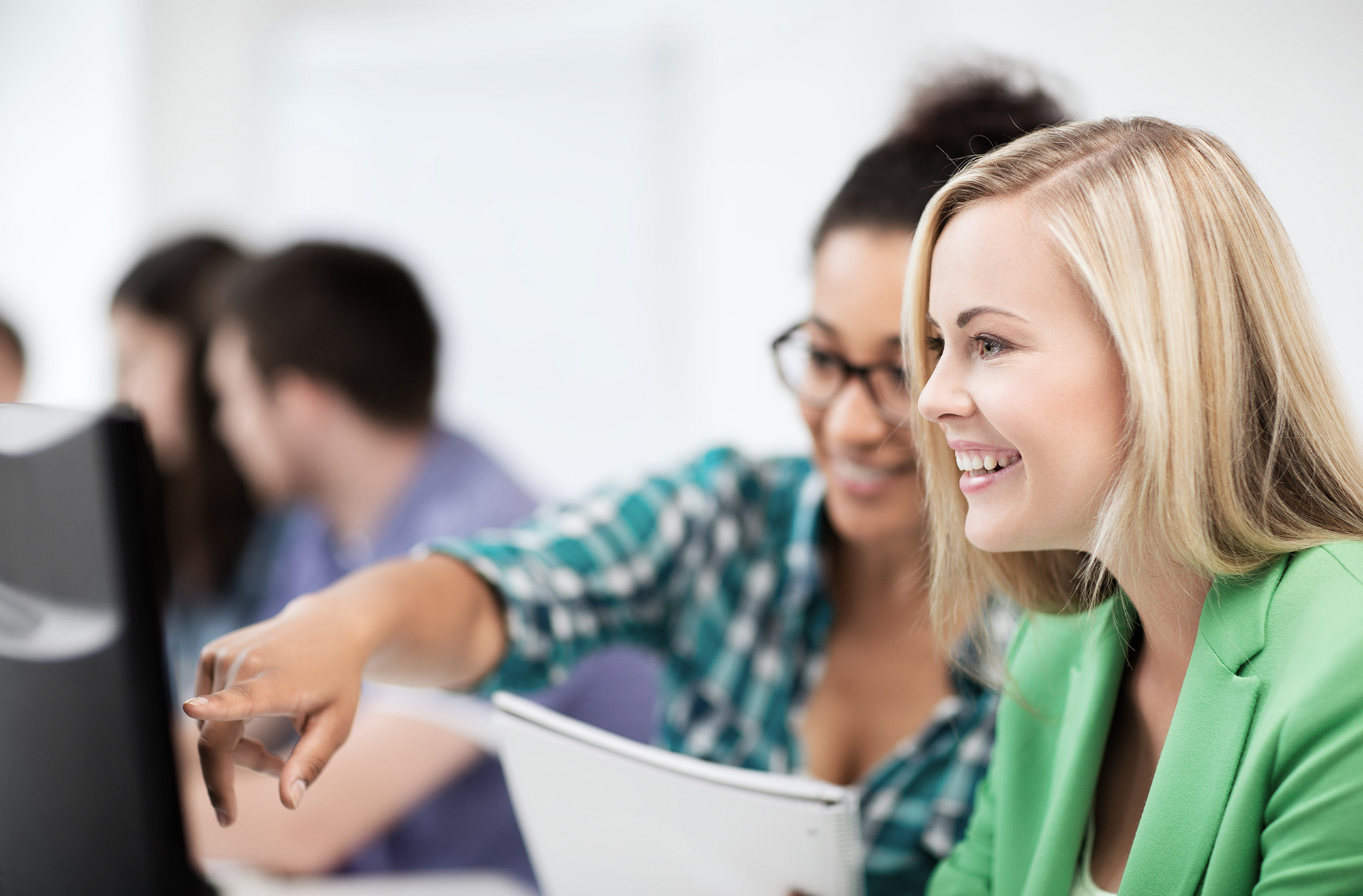 One woman smiles and looks at a computer monitor while another points to the monitor.