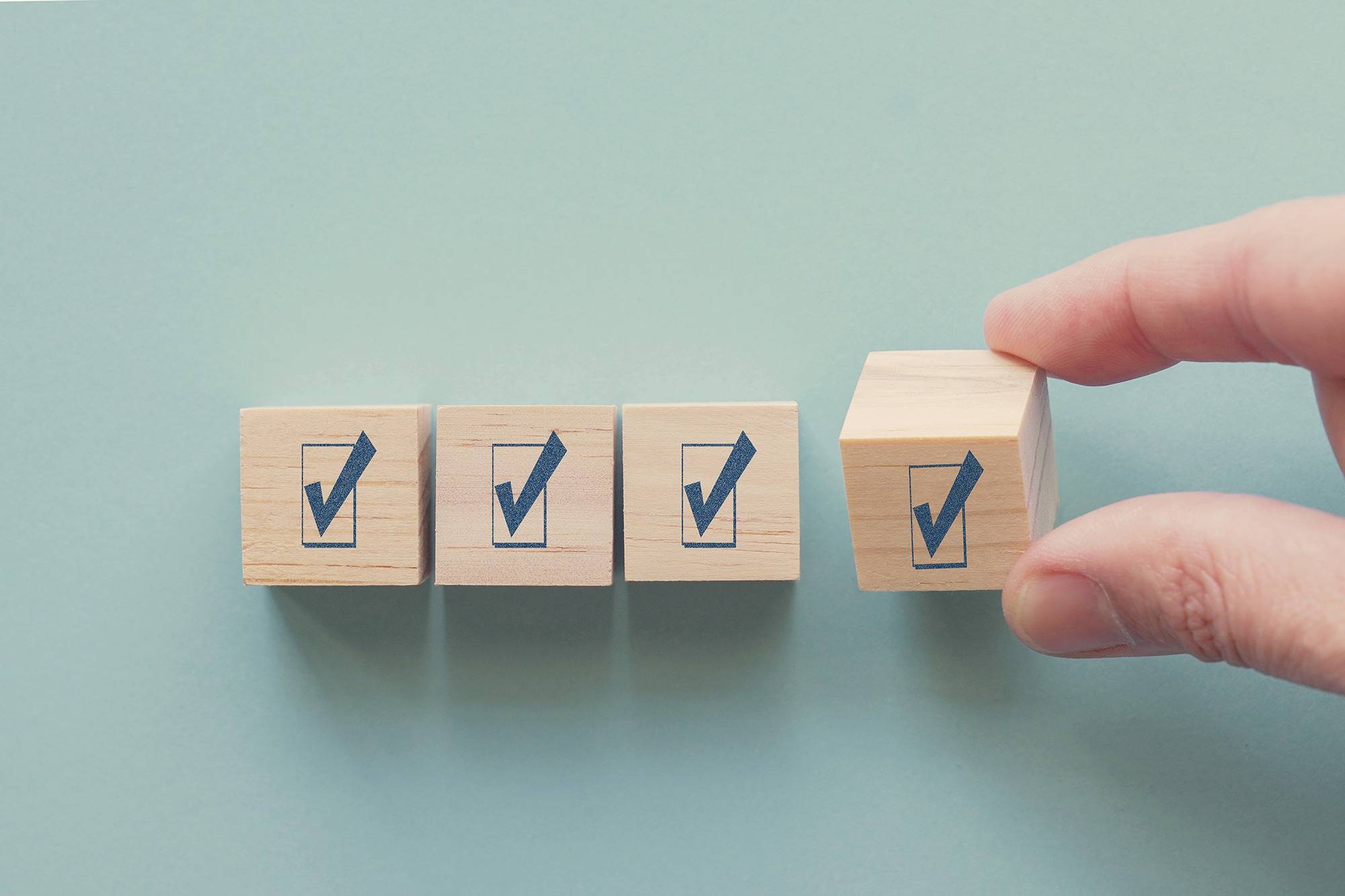 Four wooden blocks on a blue background. Each has a check mark on it. A hand is placing the final block in line with the others.