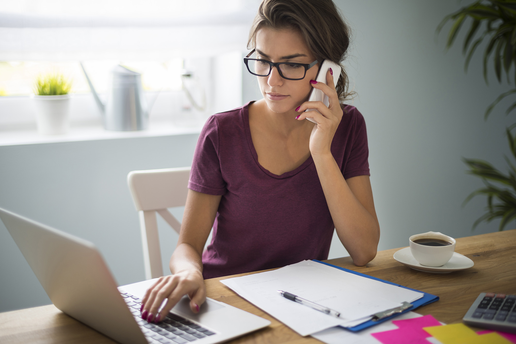 A woman uses a laptop while holding a phone to her ear. Papers are spread on the table in front of her.