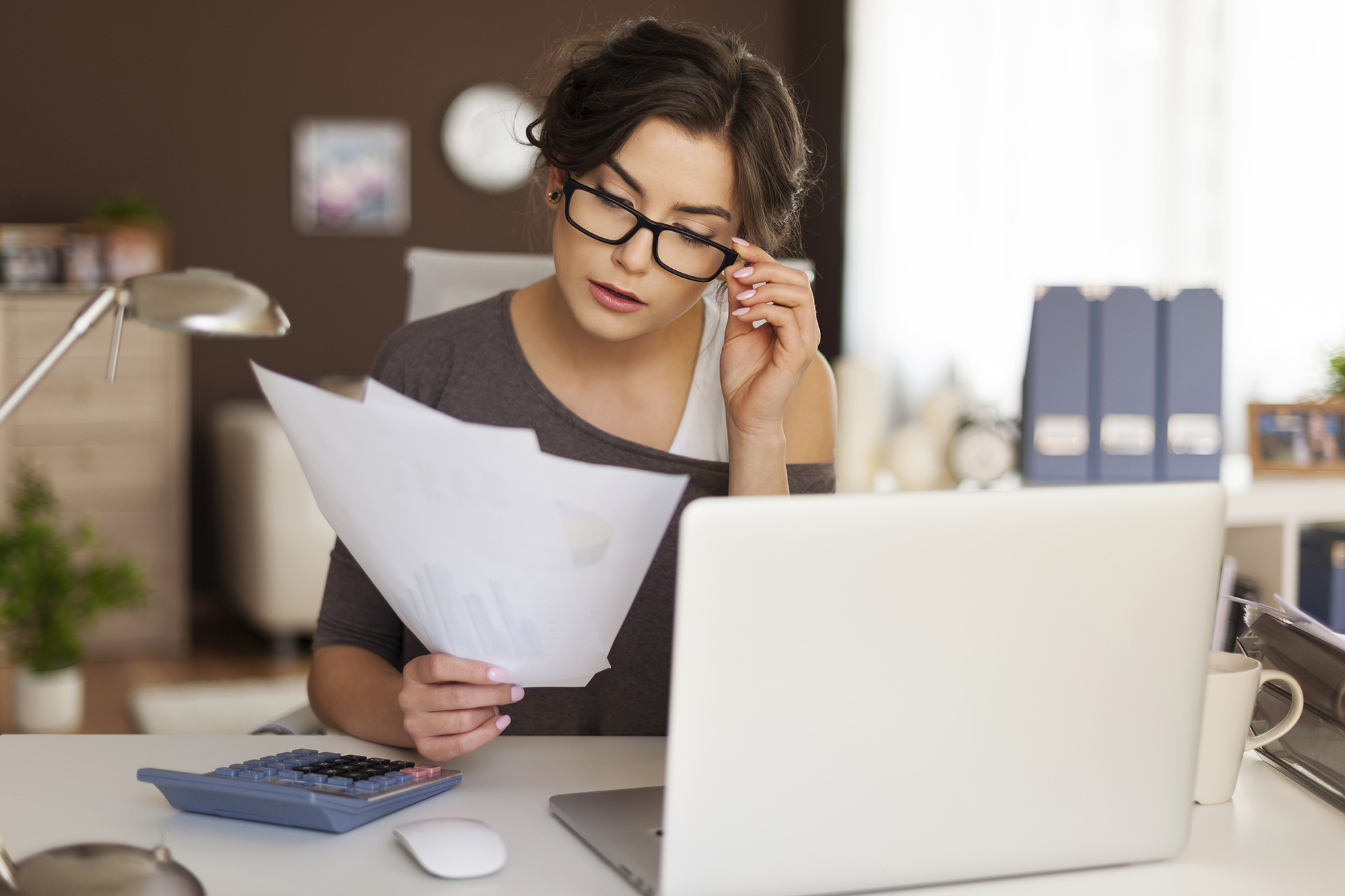 A woman in glasses reads paperwork she is holding. An open laptop and calculator sit on the table in front of her.